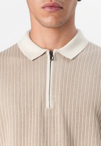 Beige ribbetty pullover med en zippered hals og en krave i cremefarve. Stoffet ser blødt ud med en lodret stribet tekstur.