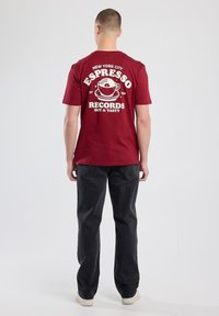 Maroon cotton t-shirt featuring a large white graphic of a coffee cup and text. Paired with dark gray jeans and light-colored sneakers.