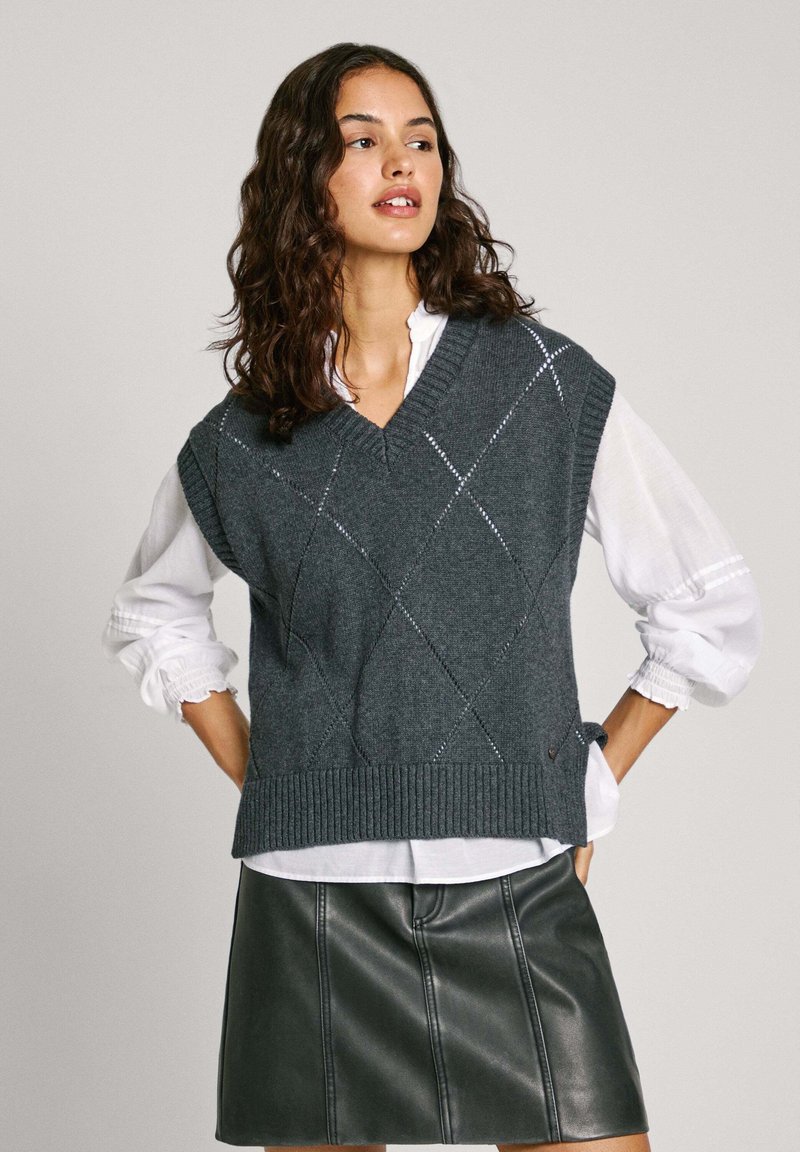 Gilet grigio a maglia con scollo a V, caratterizzato da un motivo a diamante e orlo a coste, indossato sopra una camicia bianca con maniche a sbuffo e una gonna nera in pelle.