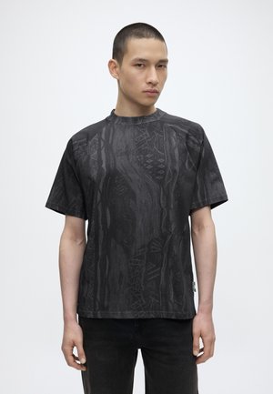 Junger Mann mit kurzen Haaren, der ein schwarzes, gemustertes kurzärmeliges Hemd und schwarze Hose vor einem weißen Hintergrund trägt.