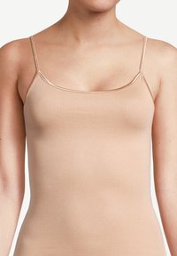 Woman wearing a beige, sleeveless, fitted camisole top with thin spaghetti straps against a plain white background.