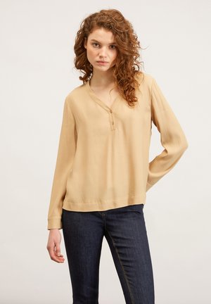 Beige blouse met lange mouwen, een V-hals en knoopdetails, gemaakt van een lichtgewicht stof. Gecombineerd met donkergekleurde jeans, minimalistisch ontwerp.