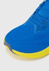 Blå atletiske skooverdel med mesh-struktur, med gul såle og grå detaljer. Designet med lisser og strømlinjeformet form.