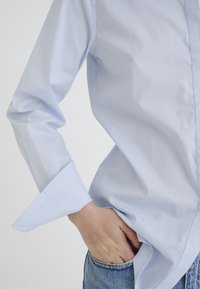 Light blue button-up shirt with long sleeves. Soft, smooth texture and pointed cuffs. Paired with denim jeans, hand positioned in pocket.