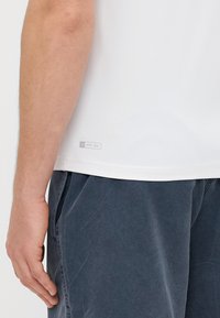 Biała koszulka UPF 50+ o gładkiej fakturze i dopasowanym kroju, połączona z ciemnoszarymi spodniami z bocznymi kieszeniami i miękkim, zużytym wyglądem.