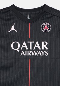 Juoda futbolo marškinėliai su vertikalią tekstūra, raudona juosta ir baltais užrašais. Priekyje yra Jordan logotipas ir „Paris Saint-Germain“ ženklelis.
