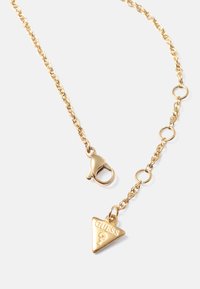 Colar de corrente em tom dourado com fecho tipo lagosta. Em destaque, uma pendente triangular gravada com "GUESS" e um ponto de interrogação.