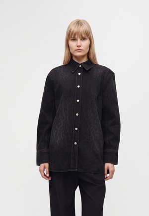 Ung kvinde med glat blondt hår iført en sort skjorte med knapper med subtilt leopardprint og sorte bukser, stående mod en ensfarvet baggrund.