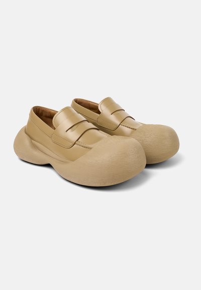 Zapatos de cuero marrón claro tipo slip-on con suelas de goma texturizadas y redondeadas, y un diseño minimalista suave que presenta dos acentos de correa en la parte superior.