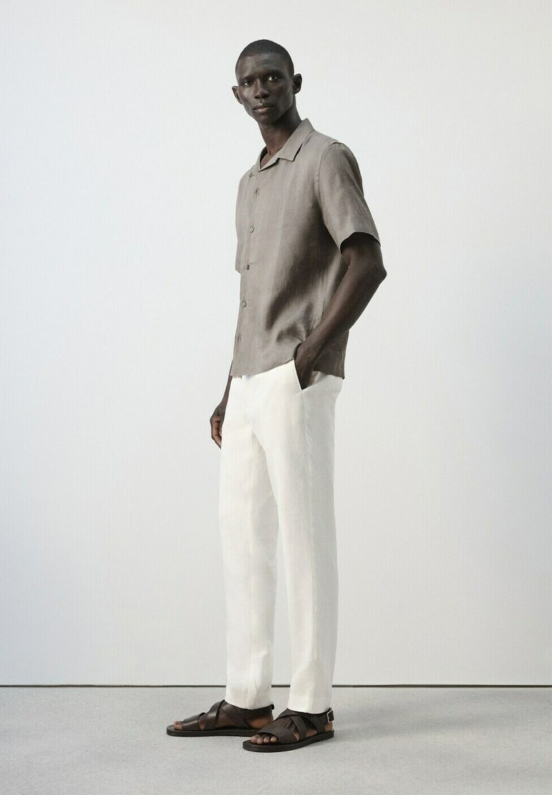 Homme debout les mains dans les poches, portant une chemise grise à manches courtes boutonnée, un pantalon blanc et des sandales marron foncé sur un sol gris.