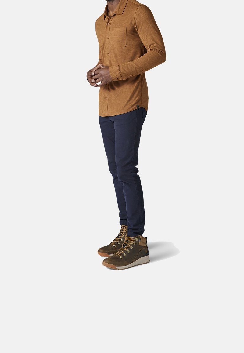 Camicia marrone a maniche lunghe con taschino, abbinata a pantaloni slim-fit blu navy e scarponi da trekking marroni con lacci dorati e suole in gomma.