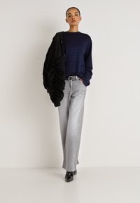 Ilgas rankovėmis dryžuotas marškinėlis jūreivių stiliaus, juoda surišama striukė, šviesiai pilkos plačios džinsinės kelnės ir juodi bateliai su smailia nosimi, su diržu, kuriame yra logotipas.