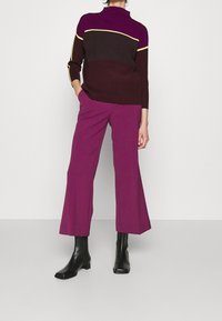 De pullover heeft een kleurblokontwerp in bordeauxrood, bruin en gele accenten; gecombineerd met paarse wijde broekspijpen en zwarte enkellaarsjes.