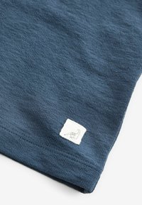 Mörkblå T-shirt med texturert tyg och en fyrkantig vit etikett som visar en dinosauriedesign i ett hörn nära fållen.