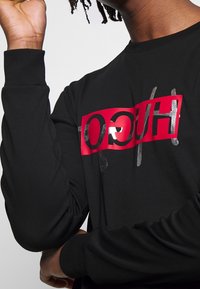 Camiseta de manga larga negra con un gráfico rojo audaz que dice "HUGO" en material reflectante plateado a lo largo del pecho. Textura de tela suave.