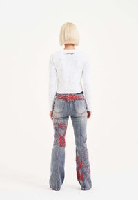 Camisa blanca de manga larga con un sutil patrón floral, combinada con unos jeans azules que tienen diseños de corazones rojos bordados y piernas acampanadas.