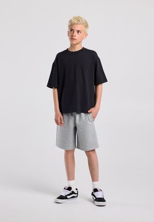 Jongen met kort blond haar draagt een loszittend zwart T-shirt, grijze shorts, witte sokken en zwarte sneakers, staand met de handen in de zakken.