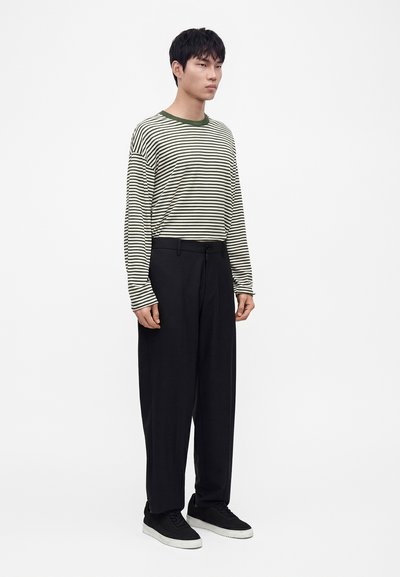 Homme portant une chemise rayée à manches longues avec un col vert, un pantalon noir à jambes larges et des baskets noires avec des semelles blanches, debout face à l’avant.