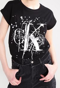 Camiseta de algodón negra de manga corta con un estampado de salpicaduras blancas y el texto 'Calvin Klein Jeans' en el centro.