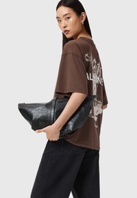 Bolso de mano negro con efecto de cocodrilo y una forma única, sostenido por una persona que lleva una camiseta oversized marrón y pantalones negros; textura suave.