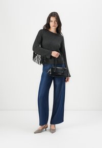 Grauer Pulli mit Fransenakzenten, blaue weite Jeans und graue spitze High Heels. Hält eine kleine schwarze Handtasche.