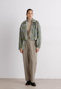 Veste courte à carreaux verts et blancs avec fermeture zippée, associée à un pantalon large beige et des chaussures foncées, sur fond neutre.