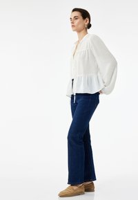 Mujer con blusa blanca de mangas largas, jeans de mezclilla azules y mocasines color beige, de pie frente a un fondo blanco, mirando hacia adelante.