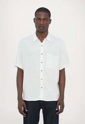 Chemise en lin blanche à manches courtes avec un devant à boutons, des boutons en bois et une poche unique sur la poitrine gauche.