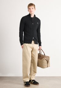 Fekete kötött zipzáras pulóver, bézs széles szárú nadrág és fekete sportcipő sárga csíkokkal. Egy barna sporttáska bőrszegélyekkel.