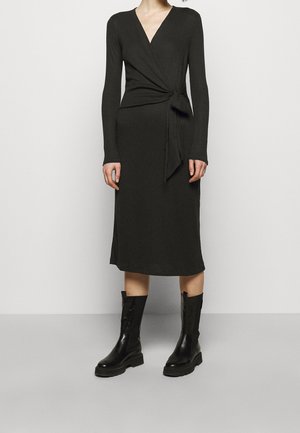 Woman wearing a long-sleeve black wrap dress with side tie, paired with mid-calf black leather boots, standing against a plain light background.