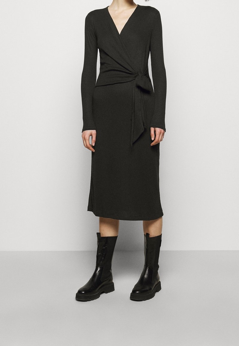 Femme portant une robe portefeuille noire à manches longues avec un lien latéral, associée à des bottes en cuir noires mi-mollet, debout devant un fond clair uni.