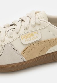 Gros plan de la basket beige Puma Palermo avec lacets blancs, détails en daim et logo Puma doré sur le panneau latéral au-dessus de la semelle.