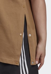 Bruine katoenen shirt met zijsplitten, vastgezet met gouden en zilveren drukknoopjes. Het heeft een rechte zoom en een gestructureerde stof.