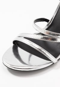 Srebrna metaliczna sandały na wysokim obcasie z krzyżującymi się paskami, gładkie wykończenie i czarne wnętrze. Otwarty czubek i smukła sylwetka.