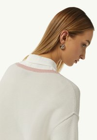 Witte gebreide trui met een textuurpatroon, voorzien van een roze accent op de halslijn. Het model draagt chunky zilveren oorbellen en een witte blouse.