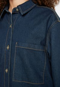 Dark denim shirt with a collar, buttoned front, and single chest pocket. Features contrasting orange stitching and a relaxed fit.