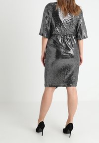 Robe à sequins argentés avec une taille cintrée, des manches courtes et un ourlet à hauteur des genoux. Présente une texture lisse et un éclat subtil. Chaussures à talons hauts noires.