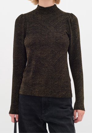 Kvinde iført en tætsiddende sort bluse med lange ærmer med gyldne glitrende bølgelinjer og sorte bukser, der holder en sort rem i den ene hånd.