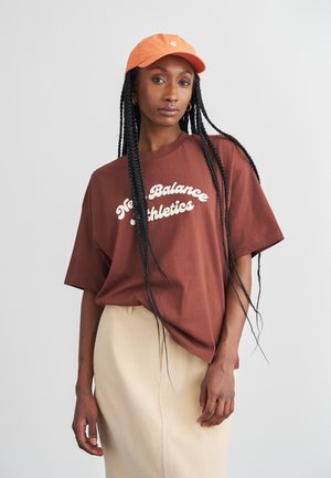 T-shirt oversized castanho com o texto branco "New Balance Athletics", combinado com uma saia creme e um boné laranja. Apresenta cabelo longo trançado e brincos.