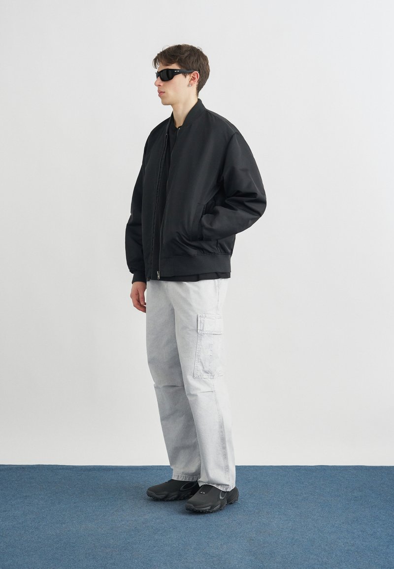 Junger Mann trägt eine schwarze Bomberjacke, weiße Cargohosen, schwarze Schuhe und schwarze Sonnenbrille, steht auf einem blauen Teppich vor einer weißen Wand.