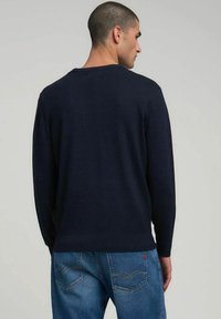 Maglione navy con maniche lunghe, scollo e orlo a coste. Indossato con jeans azzurri, caratterizzati da un motivo di cuciture sottili sulle tasche.