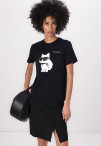 Tricou negru din bumbac cu un grafic al unei pisici albe purtând ochelari de soare. Modelul ține o geantă neagră, asortată cu o fustă neagră. Textul de pe tricou spune "KARL LAGERFELD."