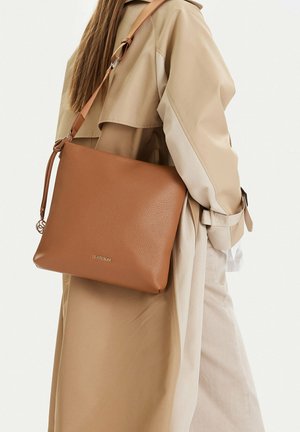 Woman wearing beige trench coat carries a large, textured brown leather shoulder bag with adjustable strap and Lasocki branding.