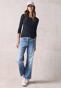 Schwarzes langärmliges Shirt kombiniert mit hellblauen weiten Jeans und weißen Sneakern. Sie trägt eine pinke Mütze und zeigt ein lässiges Outfit.