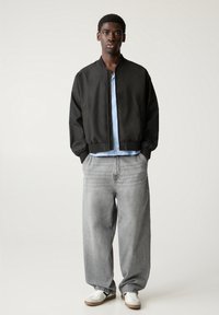 Sort letvægts bomberjakke, lyseblå skjorte med knapper, grå vide jeans og hvide sneakers med beige accenter. Simpelt design, afslappet stil.