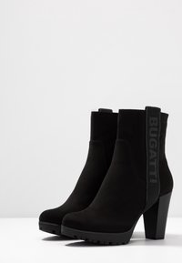 Botas negras de ante para tobillo con un tacón alto y grueso. Presentan un panel lateral con la marca "BUGATTI" en negro. Suela texturizada y resistente.
