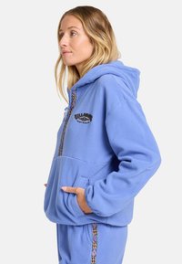 Hoodie en polaire bleu clair avec une poche avant et une fermeture éclair décorative. Présente un logo brodé et des accents à motifs le long de la fermeture éclair.