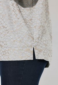 Lichtgrijs shirt met een metallic beige schubbenpatroon, met zijsplitten aan de zoom en een relaxed fit. Gecombineerd met donkere denim.