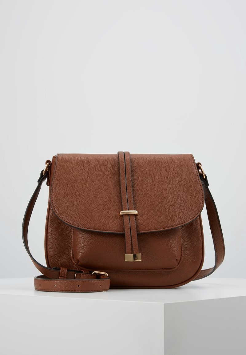 Ruskea nahkainen olkalaukku, jossa on etuläppä, kultasävyinen kiinnitys, säädettävä hihna ja kivipinta, asetettuna valkoiselle jalustalle harmaata taustaa vasten.