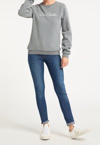 Grijze sweatshirt met geborduurd "Drei Master" logo, gecombineerd met slim-fit blauwe jeans en witte sneakers. Zachte textuur, ronde halslijn, lange mouwen.